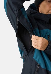 Man wearing black beanie unzips underarm ventilation zipper on blue and black waterproof jacket.