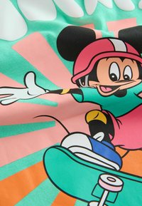 Mickey Mouse draagt een roze helm en een groenblauw shirt, skateboardend met uitgestrekte armen tegen een levendige roze en groenblauwe achtergrond.