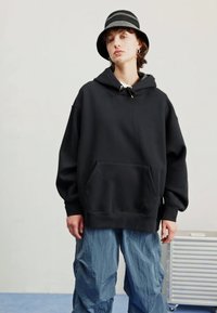 Sudadera negra de gran tamaño con bolsillo canguro y capucha con cordón, combinada con pantalones azules sueltos. La tela parece suave y texturizada.