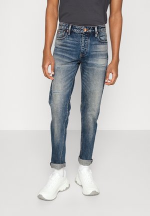 Osoba w podwiniętych wyblakłych niebieskich dżinsach, białych masywnych trampkach i ciemnoszarej koszuli stojąca na jednolitym tle.