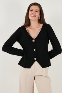 Junge Frau mit langem braunem Haar lächelt, trägt eine schwarze V-Ausschnitt-Strickjacke mit goldenen Sonnen- und runden Knöpfen sowie beige hochgeschnittene Hosen.