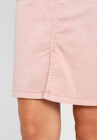 Ljusrosa bomullsskjorta med rak skärning, med synlig sömnad längs fållen och sömmen på sidan. Slät textur, knälång design.