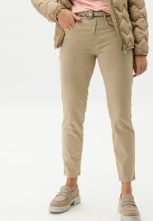Personne portant un pantalon beige slim, une ceinture beige avec boucle double anneau, une doudoune marron, une chemise rayée et des mocassins beiges.