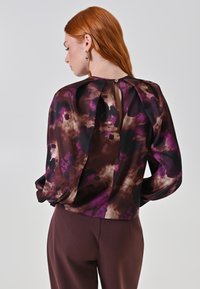 Persona dai capelli rossi che indossa una camicetta a maniche lunghe con un motivo astratto in viola, nero e beige, in piedi di spalle rispetto alla camera su uno sfondo semplice.