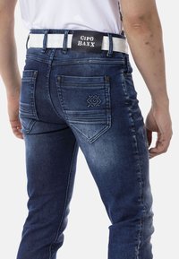 Jeans en denim bleu foncé avec une coupe ajustée, poches arrière avec des coutures décoratives et une ceinture blanche contrastante avec logo.