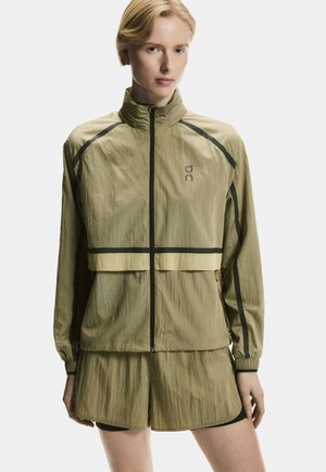 Plavolaska ženska, oblečena v olivno zeleno lahko jakno z zadrgo in ujemajoče hlače, stoji pred belim ozadjem.