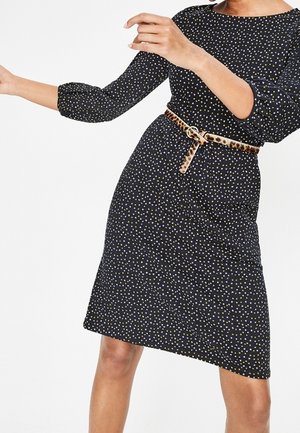 Femme portant une robe noire à petits pois blancs et jaunes, manches trois-quarts, et une ceinture imprimé léopard à la taille.