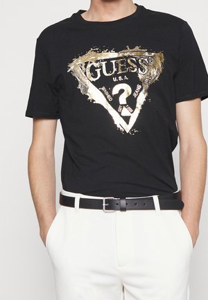 Homme portant un T-shirt noir avec le logo Guess métallique doré, pantalon blanc, ceinture noire, les mains dans les poches, debout devant un fond blanc.