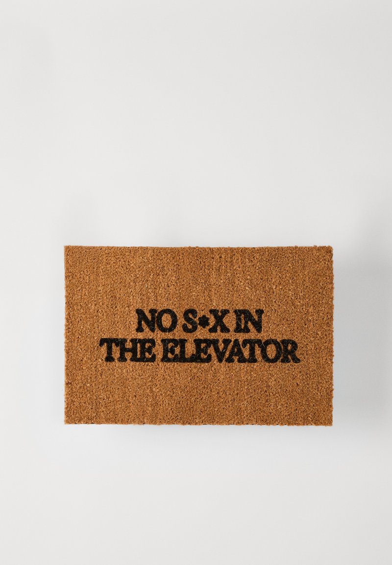 Korķa durvju paklājs ar izteiksmīgu melnu tekstu "NEVEICIET SEKSU LIFTĀ," taisnstūrveida forma, teksturēta virsma, dabīga brūna krāsa.