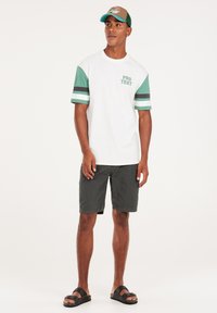Witte t-shirt met groen en zwarte gestreepte mouwen en "PRO TEST" op de voorkant gedrukt. Gecombineerd met donkere shorts en zwarte sandalen.