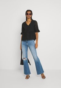 Top de crochê preto com mangas curtas em formato de sino, decote em V, barra com ondas; combinado com jeans flare azul claro com detalhes desgastados. Bolsa preta e óculos de sol.