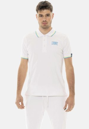 Junger Mann steht, trägt ein weißes Poloshirt mit blau-gelbem Rand und "LEONE"-Logo, kombiniert mit weißen Hosen.