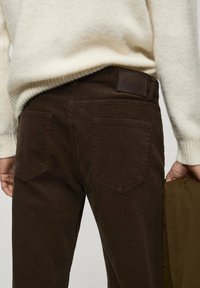 Bruine corduroy broek heeft een gestructureerd oppervlak, achterzakken met schuine kleppen en een donkerleren patch op de tailleband.