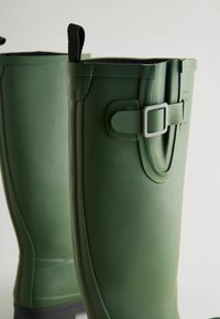 Groene rubberen regenlaarzen met een gladde textuur, voorzien van een zijgesp en een zwarte trekband aan de bovenkant voor gemakkelijke bediening.