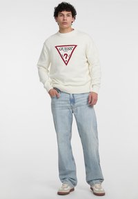 Hvid strikket sweater med et rødt trekantet logo med "?". Kombination med lyseblå baggy jeans og beige sneakers med hvide accenter.