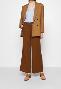 Mujer vistiendo pantalones amplios marrones con un cinturón a juego, un blazer camel de doble botonadura, una blusa a rayas azul claro y sandalias marrones de punta abierta.