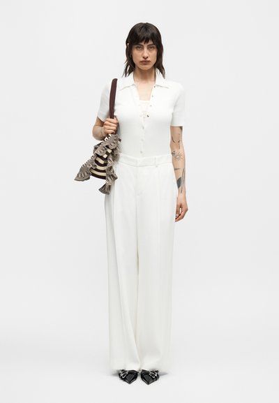 Γυναίκα με σκούρα μαλλιά, που φοράει λευκή κοντομάνικη μπλούζα με κουμπιά, φαρδιά λευκά παντελόνια, κρατάει ριγέ τσάντα με βολάν και μαύρα μυτερά παπούτσια.