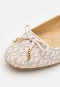 Chaussure plate beige avec texture à motifs, nœud noué sur le dessus et breloques métalliques dorées attachées aux extrémités du nœud sur fond blanc.