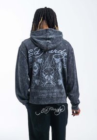 Sudadera negra con estampado de tie-dye, con un gráfico de calavera y llama en la parte posterior. Presenta el logo de "Ed Hardy" y detalles de dirección. Mangas largas y corte relajado.
