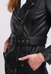 Černá kožená bunda s páskem v pase, zapínáním na zip a velkými kapsami. Obsahuje klopový límec a stříbrné kovové doplňky.