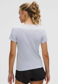 Vrouw met een krullende paardenstaart, gekleed in een lichtgrijs sportshirt met korte mouwen en zwarte hardloopshorts, gezien vanuit de achterkant.
