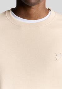 Beige sweatshirt met een zachte textuur en ribbelhals, voorzien van een subtiele geborduurde vogel logo op de linkerkant van de borst.