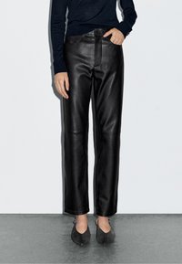 Schwarze Lederhose mit geradem Bein, glatter Textur und vorderen Taschen. Kombiniert mit dunkelgrauen, spitzen Ballerinas.