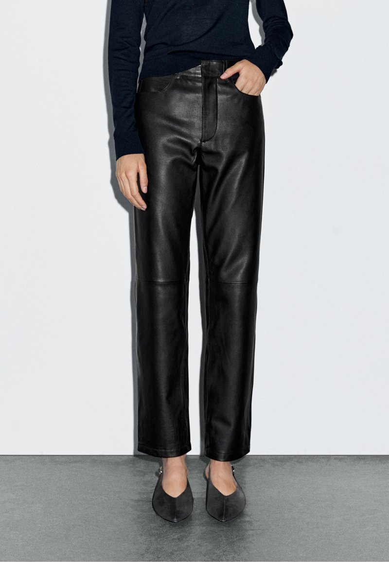 Schwarze Lederhose mit geradem Bein, glatter Textur und vorderen Taschen. Kombiniert mit dunkelgrauen, spitzen Ballerinas.