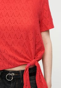 Κόκκινο κοντομάνικο πλεκτό τοπ με σχέδιο από μικρές οπές, δεμένο με κόμπο πάνω από μαύρο τζιν με ζώνη με στρογγυλή ασημένια αγκράφα.