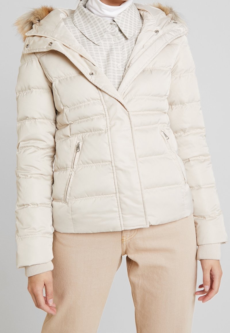 Giacca leggera tipo puffer color crema con cuciture orizzontali, cappuccio staccabile con bordo in pelliccia sintetica e due tasche laterali con zip.