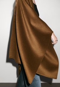 Brun ullponcho som hänger smidigt över axeln, med en mjuk textur och vinklad skärning, kompletterad med åtsittande blå denim.