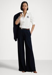 Bela srajca s pokončnimi gumbi z mornarsko modrim logotipom, visokim pasom in širokimi hlačami v mornarsko modri barvi ter rjavim pasom. Model drži mornarsko modri blazer, na nevtralnem ozadju.