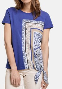 Woman wearing a short-sleeve blue shirt with geometric patterned scarf print and a tied knot on the right side, paired with light beige pants.