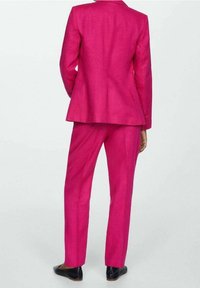 Felroze pakset bestaande uit een getailleerde blazer en bijpassende broek, beide met een gladde textuur en een aansluitend silhouet. Zwarte schoenen gedragen.