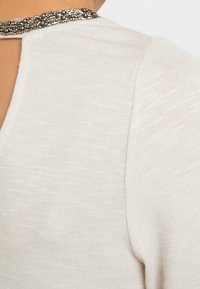 Haut beige clair en tissu texturé, avec un dos à trou de serrure et un accent décoratif au col en perles. Manches courtes et coupe décontractée.