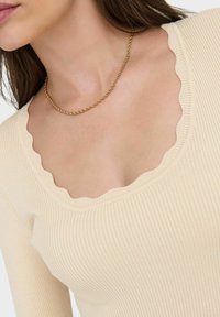Licht beige geribbelde lange mouwen top met een gegolfde halslijn. De stof is zacht en gestructureerd, aangevuld met een delicate gouden ketting.