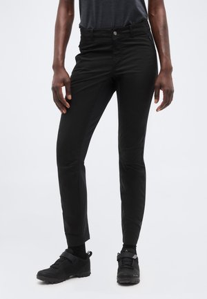 Persoană purtând pantaloni negri conici, șosete negre și pantofi sport negri, stând pe un fundal simplu, deschis la culoare.