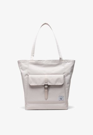 Beige tote bag med dobbel skulderrem, frontlomme med klap sikret af en spænde, og et lille rektangulært logo-patch nederst til højre.