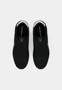 Black sneakers with a smooth, textured upper, featuring tonal stitching, laces, and a white accent on the heel. Brand logo visible on tongue.