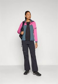 Donna con giacca rosa e verde acqua, pantaloni neri, scarponi da trekking e zaino nero, in piedi davanti a uno sfondo semplice.