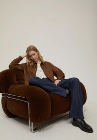 Poltrona in velluto tuftato marrone con finiture in cromo, caratterizzata da braccioli arrotondati e un design accogliente. Una persona è seduta con nonchalance in jeans scuri e stivali.