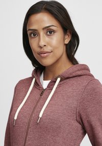 Burgunderfarget zip-up hettegenser med snor og kremfargede detaljer, teksturert stoff og gullfargede detaljer. Hvit t-skjorte synlig under.