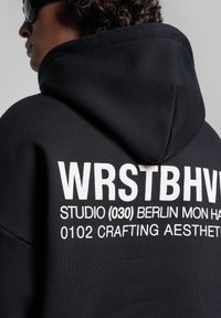 Felpa nera con cappuccio, caratterizzata da una grande stampa bianca sul retro con la scritta "WRSTBHVR" e ulteriori informazioni sul marchio. Tessuto morbido e testurizzato, design con cappuccio.