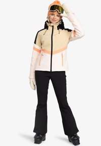 Bežová a ružová lyžiarska bunda s čiernymi akcentmi, s vreckami na zips a farebným blokovaním. Čierne lyžiarske nohavice a rukavice dopĺňajú celý outfit.