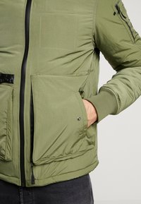 Main gauche dans la poche à rabat ouverte d'une veste matelassée vert olive avec une fermeture éclair noire et un poignet côtelé sur la manche droite.