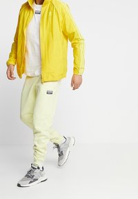 Žlutá lehká bunda s bílými pruhy, bílý tričko, světle žluté tepláky a šedé tenisky s černými akcenty.