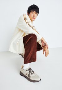 Beiga sneakers med mesh- och texturdetaljer, ihop med bruna kordflamsbyxor och en krämfärgad skjorta med knappar som har bruna ränder.