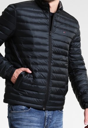 Homme portant une veste matelassée noire zippée avec poches latérales et un jean bleu, main dans la poche.