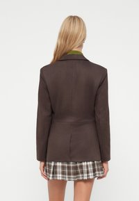 Bruine op maat gemaakte blazer met een gladde textuur, enkele knoopsluiting en een getailleerde taille. Draag het over een geruite rok met een open kraag.