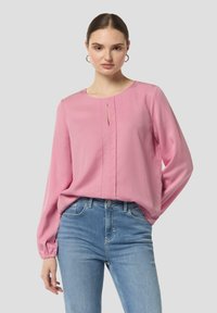 Langärmlige pinke Bluse mit rundem Ausschnitt, zentralem Falten-Detail und elastischen Ärmelabschlüssen, kombiniert mit hellblauen Jeans.